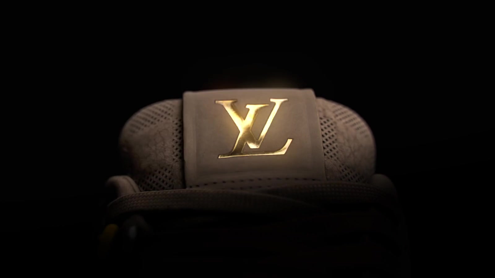 A close-up of a taupe sneaker’s tongue, gold monogram, woven fabric, and laces under studio lighting.
